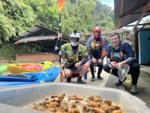 Rafting and Chonta Curo Three kayakers in helmets and PFDs pose next to a tray of grilled chonta curo grubs, with colorful kayaks and lush green forest in the background.