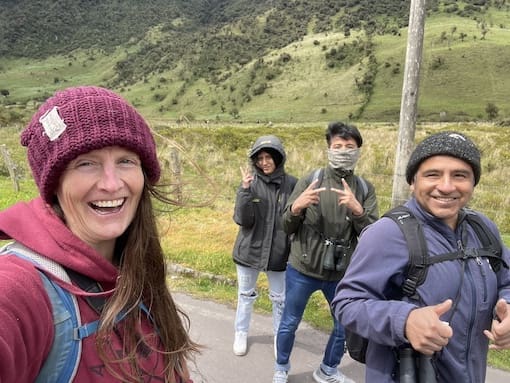 Four friends bundled up in cozy hats and jackets, smiling and flashing peace signs on a roadside walk through Ecuador’s highlands, with rolling green hills in the background.